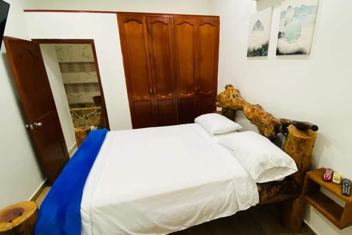 A bedroom with a wooden bed frame, a wardrobe, a small bedside table, and a door leading to another room.