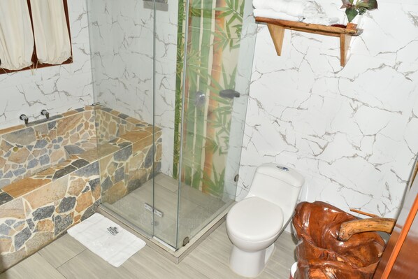 A bathroom with a stone bathtub, a glass-enclosed shower, a toilet, and a wooden sculpture. - eco hostal kuima (Santa Marta)