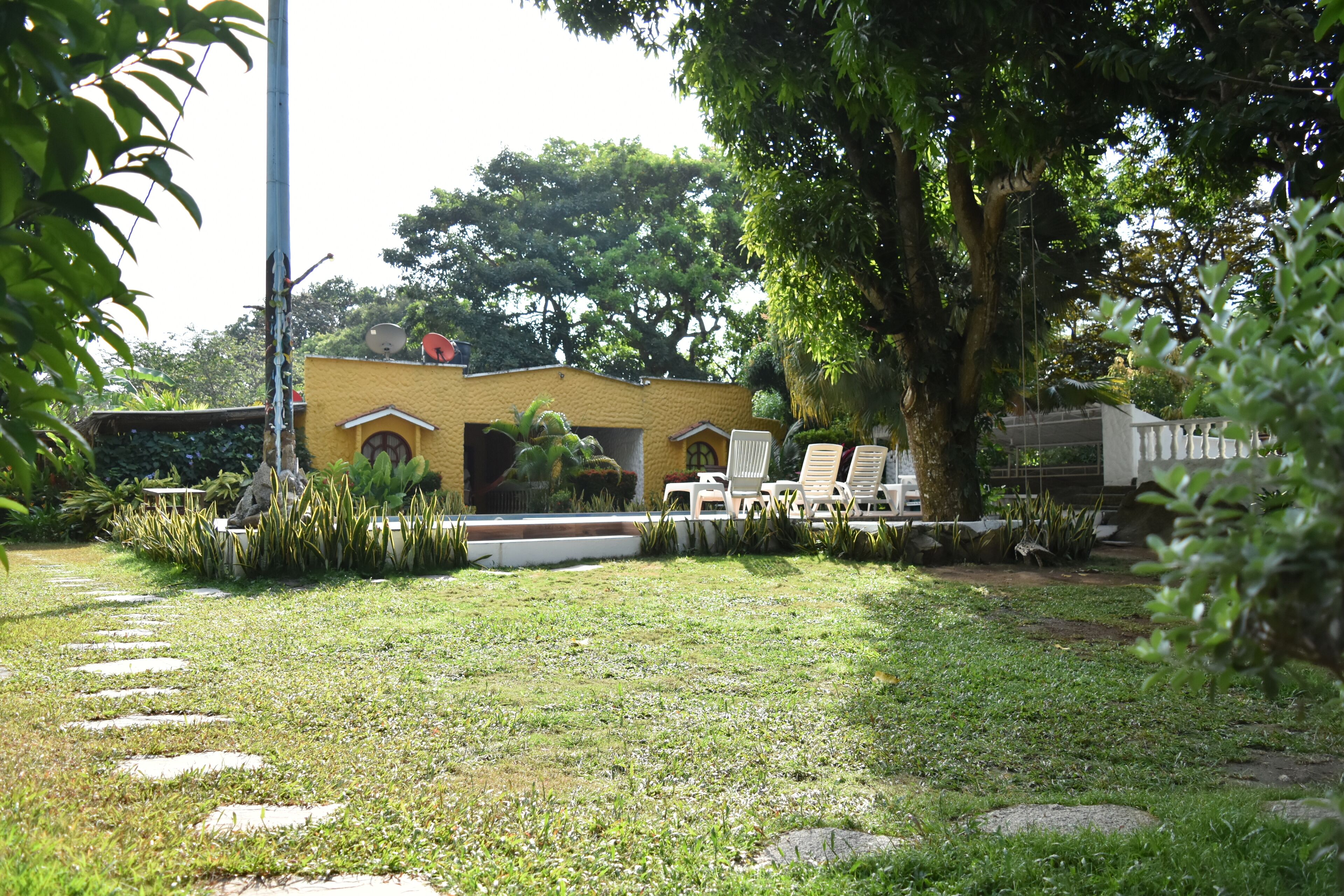 A garden with a yellow building, white chairs, and a large tree.