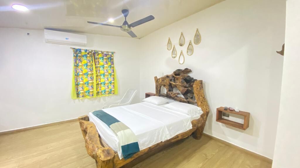 A bedroom with a wooden bed, a ceiling fan, a wall-mounted air conditioner, a window with a colorful curtain, and a wall decoration.