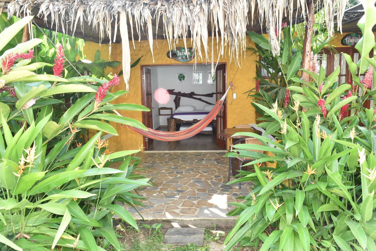 A hammock under a thatched roof, a stone-tiled entrance, and lush tropical plants.