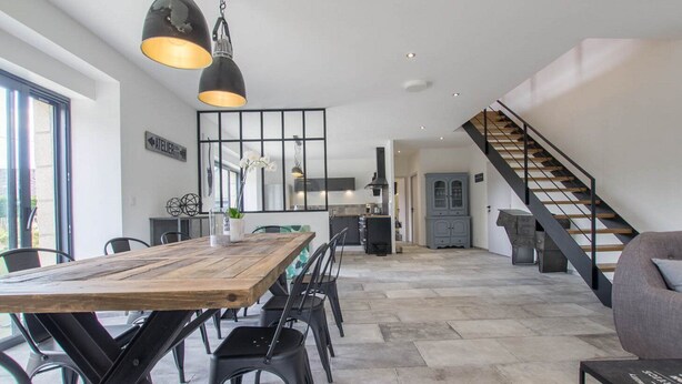 Ein geräumiger Wohnbereich mit Esstisch aus Holz, schwarzen Stühlen und Pendelleuchten. Eine Treppe führt in einen oberen Bereich, und im Hintergrund ist eine Küche erkennbar.