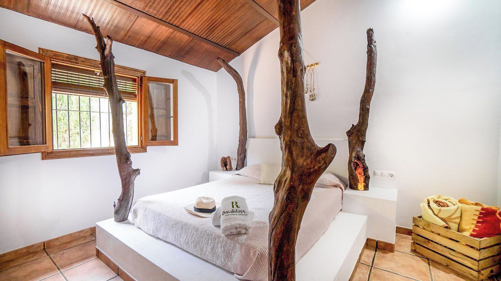 Un dormitorio con una cama grande, decoración de madera con forma de árbol, una ventana con persianas y una mesita de noche con lámpara.