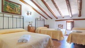 A hotel room with three beds, wooden beams on the ceiling, a painting on the wall, and a window with blinds. - Cortijo rural Majolero Castillo de Locubín by Ruralidays (Jaén)