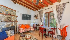 A cozy living room with a stone fireplace, a sofa with orange cushions, a dining table with chairs, and a large window with patterned curtains.