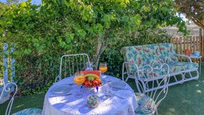 An outdoor dining area with a table set for two, surrounded by lush greenery and a wooden fence.