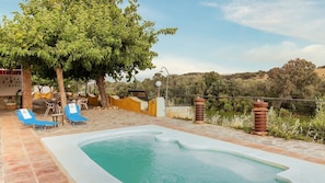 A swimming pool with blue lounge chairs, a tree, and a view of hills.