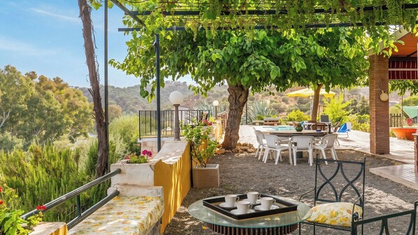 An outdoor patio with a table and chairs, a pergola with vines, and a view of the surrounding landscape.