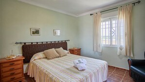 Un dormitorio con cama, mesita de noche, cómoda, ventana con cortinas y una silla.