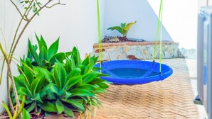 Un trampolín azul sobre un patio empedrado de ladrillo, con plantas verdes y una pequeña palmera.