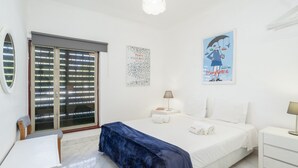 A hotel room with a bed, a desk, a chair, a window with blinds, a wall with a poster, and a nightstand with a lamp. - Vilamoura Aldeia do Mar by Hopstays | Pool (Vilamoura)