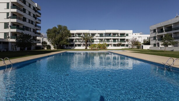 A rectangular swimming pool with clear blue water, surrounded by a paved deck. In the background, there are multi-story buildings with balconies and windows. - Vilamoura Aldeia do Mar by Hopstays | Pool (Vilamoura)