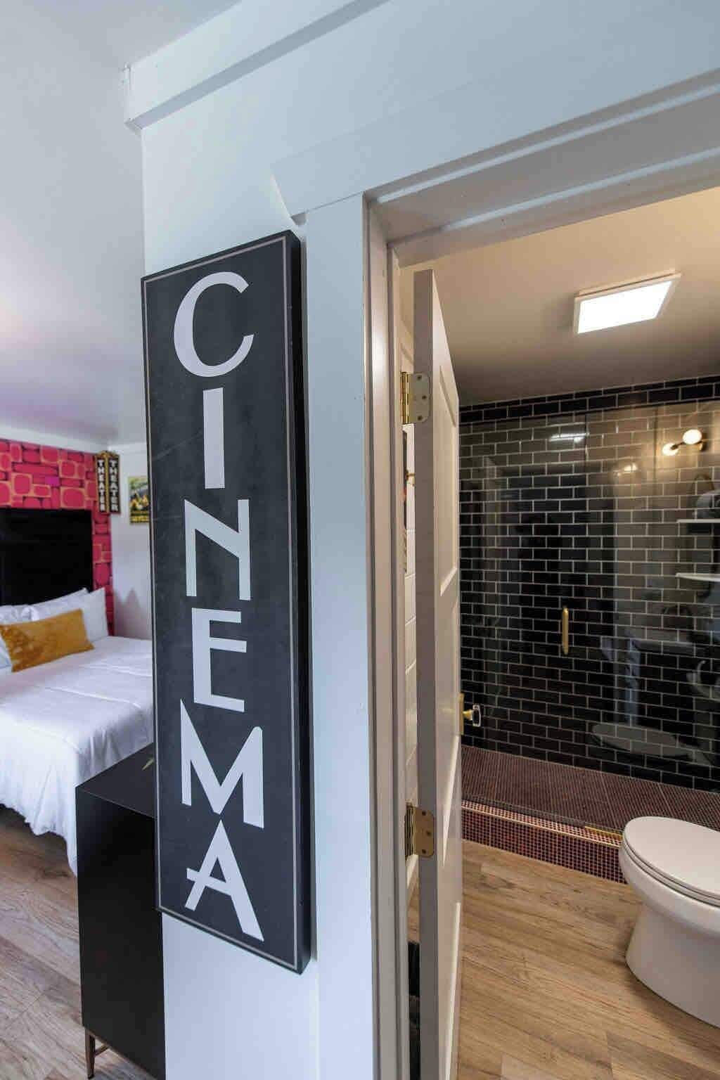 A hotel room with a bed, a bathroom with a black tiled wall, and a sign that reads 'CINEMA'.