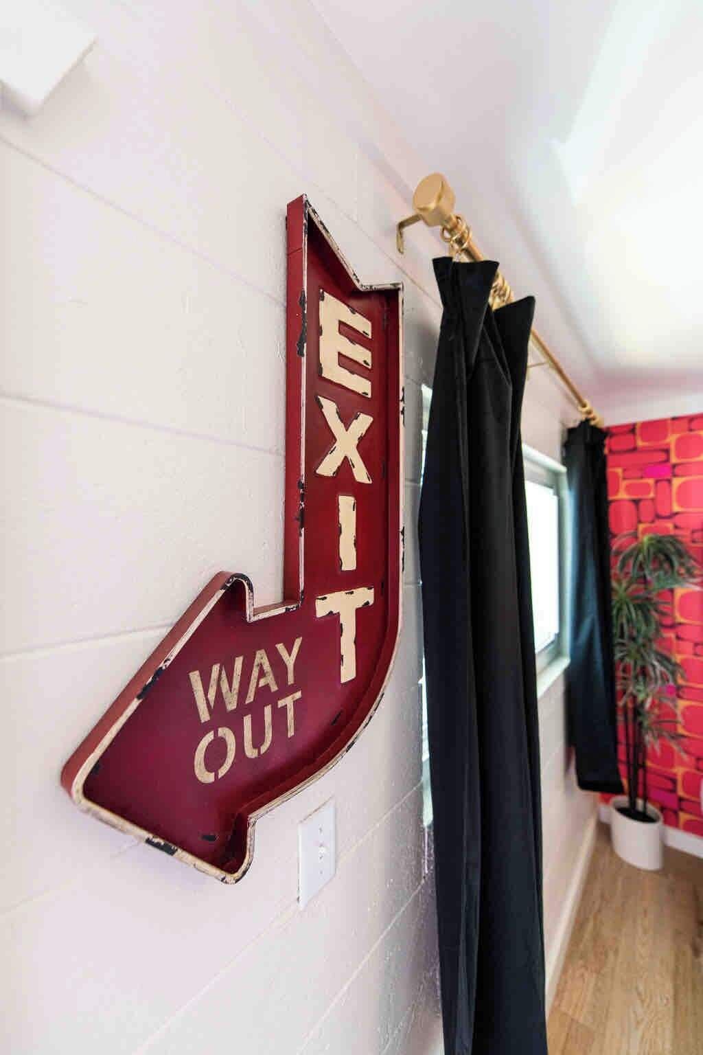 Un panneau fixé au mur avec les mots « SORTIE » et « DIRECTION DE LA SORTIE », un rideau noir et, à l’arrière-plan, un mur rouge à motifs.
