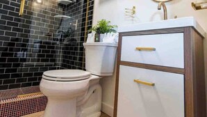 A modern bathroom with a toilet, a vanity with drawers, a mirror, and a shower area with black tiles.