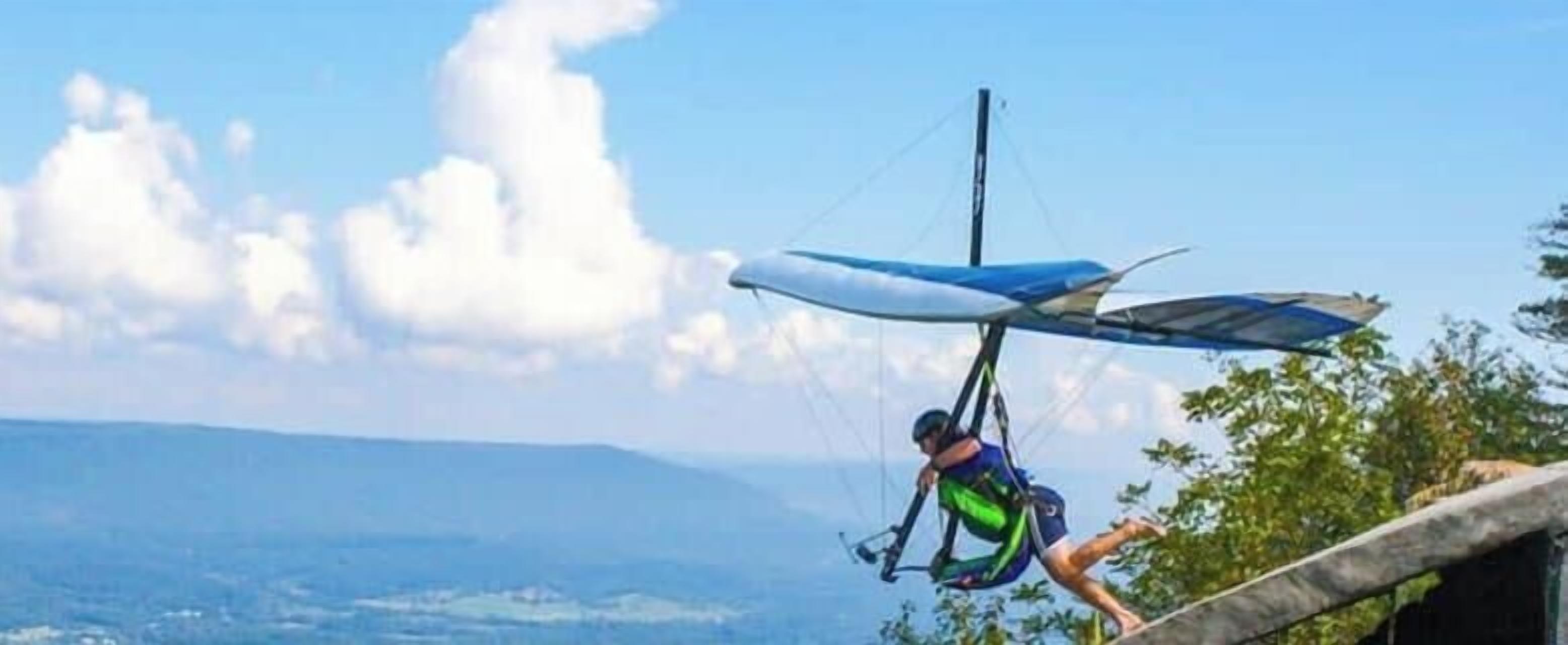 Una persona practicando parapente con un ala unida a un arnés, cielo despejado y colinas a lo lejos.