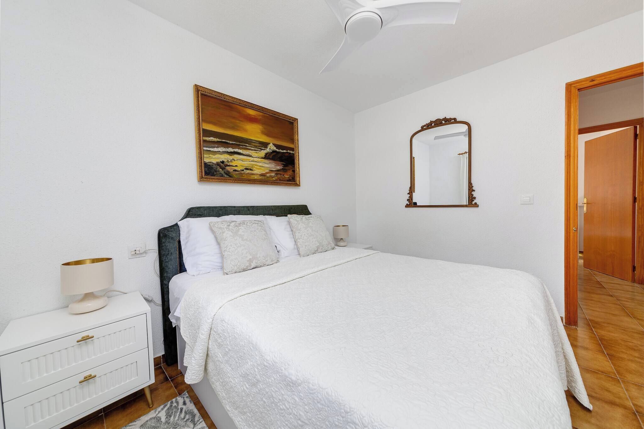 A bedroom with a bed, a nightstand, a lamp, a painting, a mirror, and a door.