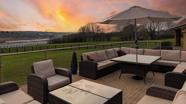 Patio esterno con mobili in vimini, un tavolo e un ombrellone, con vista su un fiume e un paesaggio verde al tramonto.