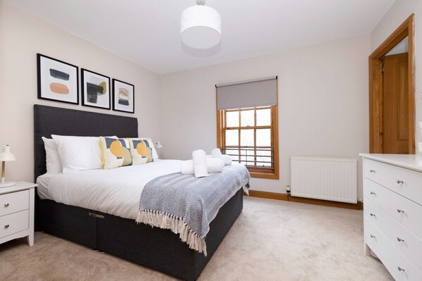 Un dormitorio con cama, mesitas de noche, cómoda, ventana con persianas y cuadros en la pared.