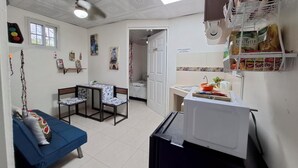 Een knus woongedeelte met een kitchenette, een zithoek met een blauwe bank en stoelen, een kleine tafel en een deur naar een andere kamer.