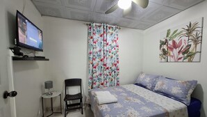 Een slaapkamer met een bed, een stoel, een klein tafeltje, een televisie en een plafondventilator.