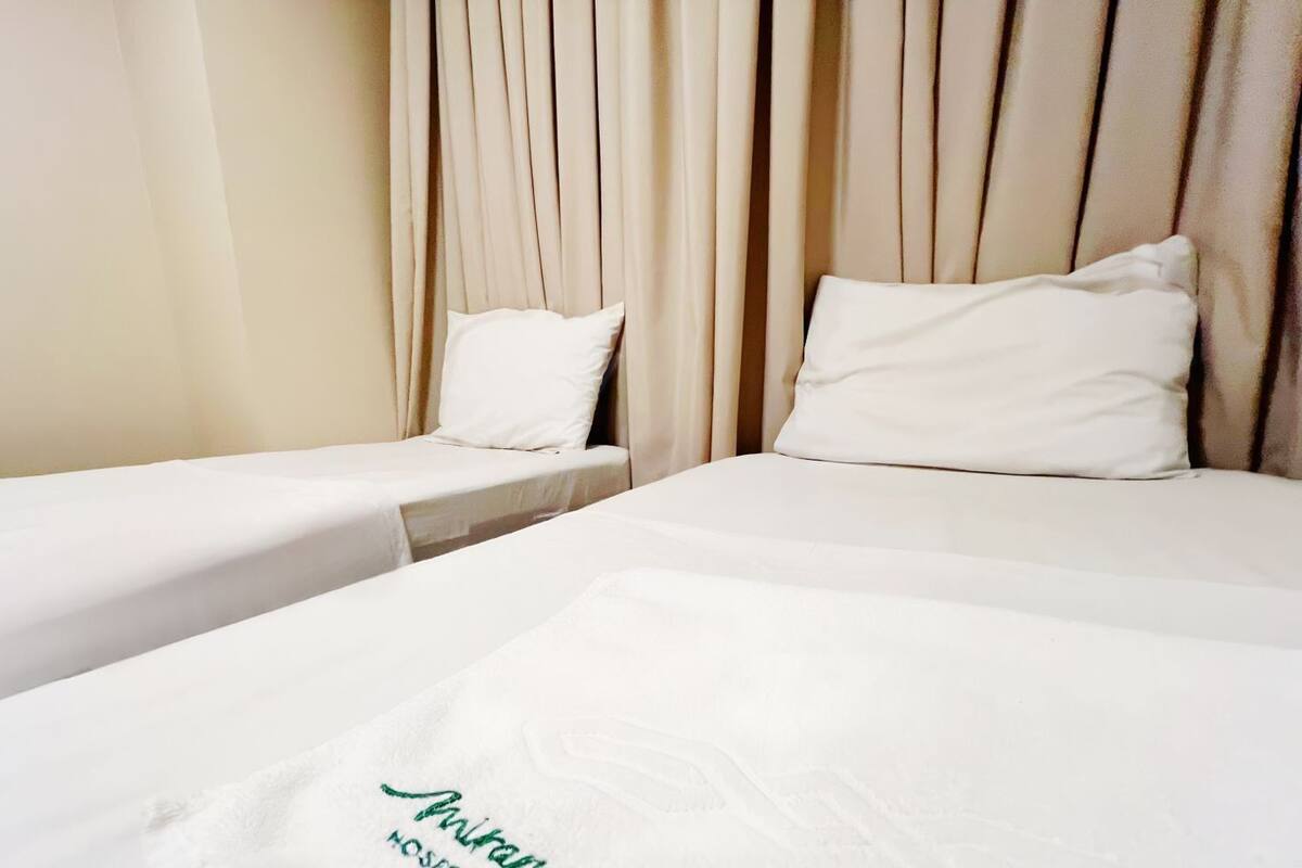 Ett hotellrum med två sängar, beige gardiner och ett vitt sängöverkast med en logotyp.