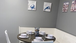Zona pranzo con tavolo rotondo apparecchiato per quattro persone, sedie bianche e una panca. Alle pareti, sopra il tavolo, si vedono opere d'arte incorniciate.