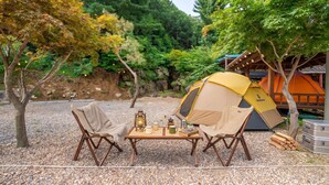 A camping setup with a tent, two chairs, a table with camping gear, and a forested backdrop. - Gapyeong Dolgungpensyeon&kaempingjang (Gapyeong)