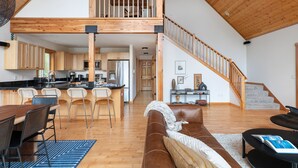 A spacious loft with a high ceiling, wooden beams, a kitchen with bar stools, a living area with a sofa and coffee table, and a staircase. - A-frame at Goss Lake (Langley)