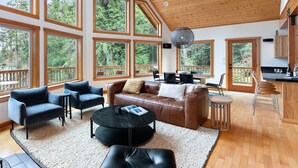 A spacious living room with large windows, a wooden ceiling, and a cozy seating area with a leather couch and chairs. - A-frame at Goss Lake (Langley)