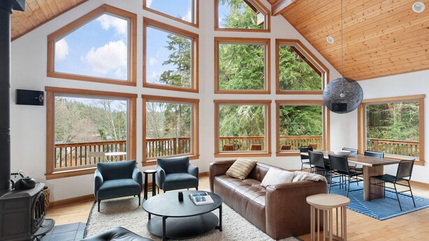A spacious living room with large windows, a fireplace, a sofa, a coffee table, and a dining area. - A-frame at Goss Lake (Langley)
