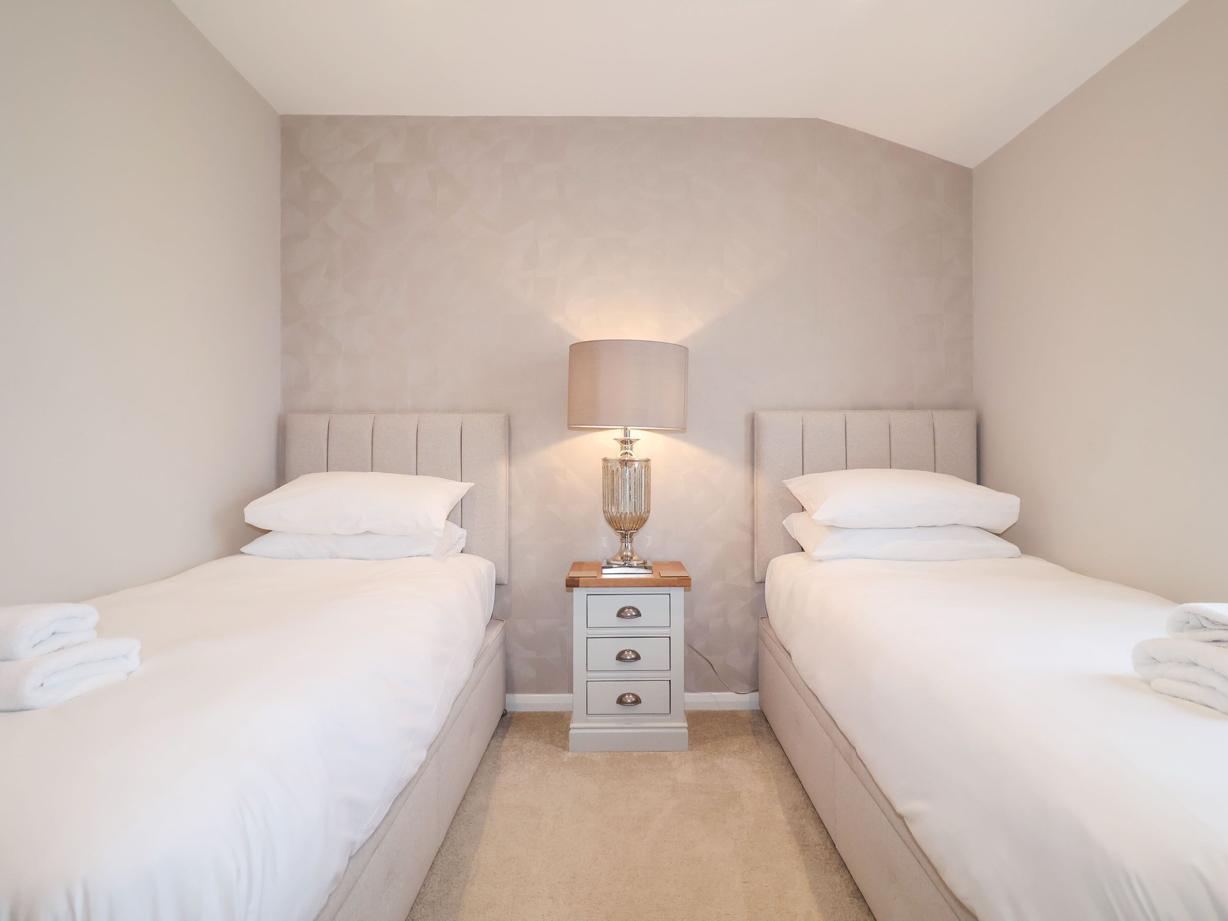 A hotel room with two beds, a nightstand with a lamp, and a plain wall.