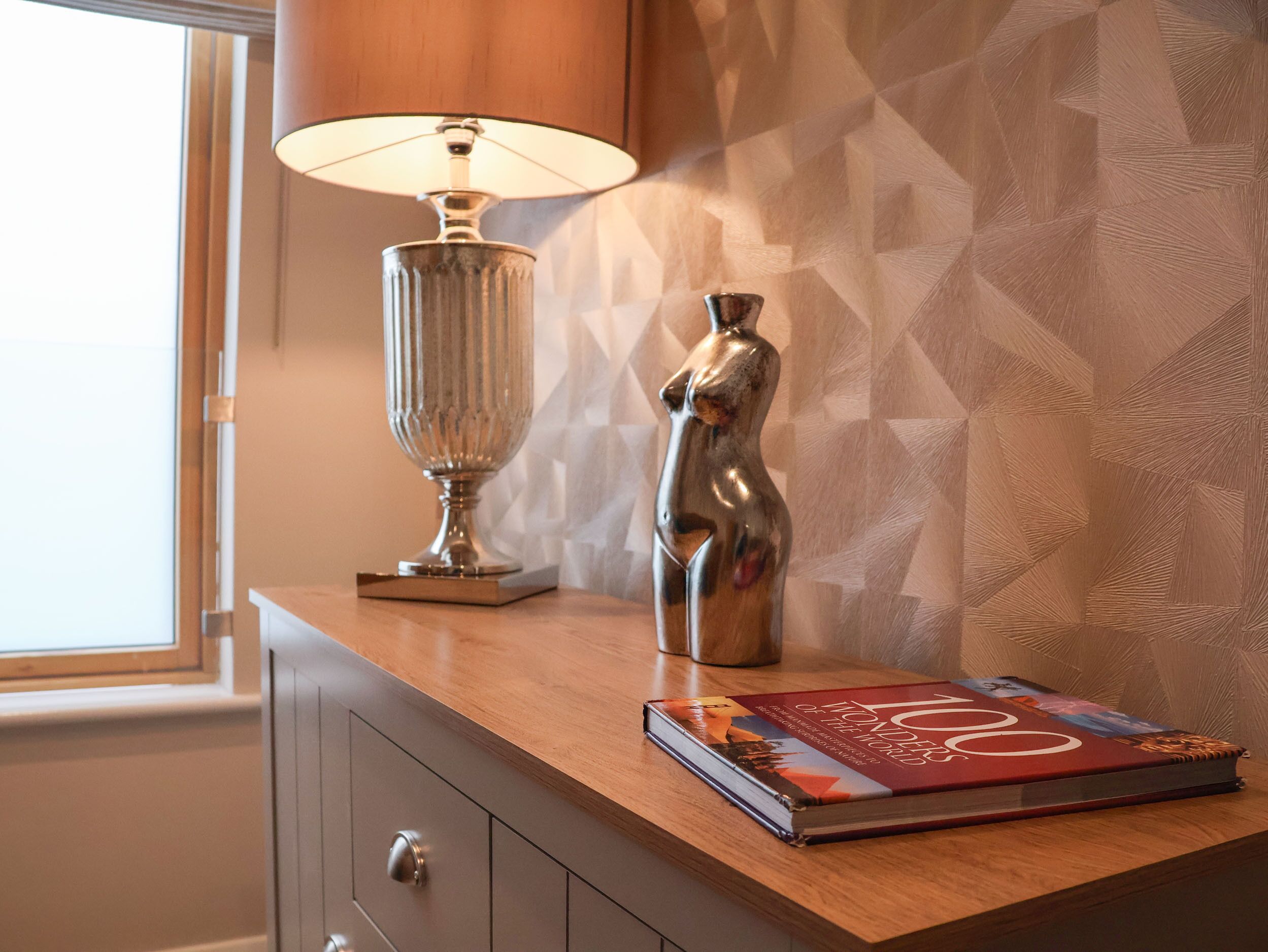 Ein Holznachttisch mit Lampe, einer Dekorationsvase und einem Buch mit dem Titel '100 Wonders of the World'.