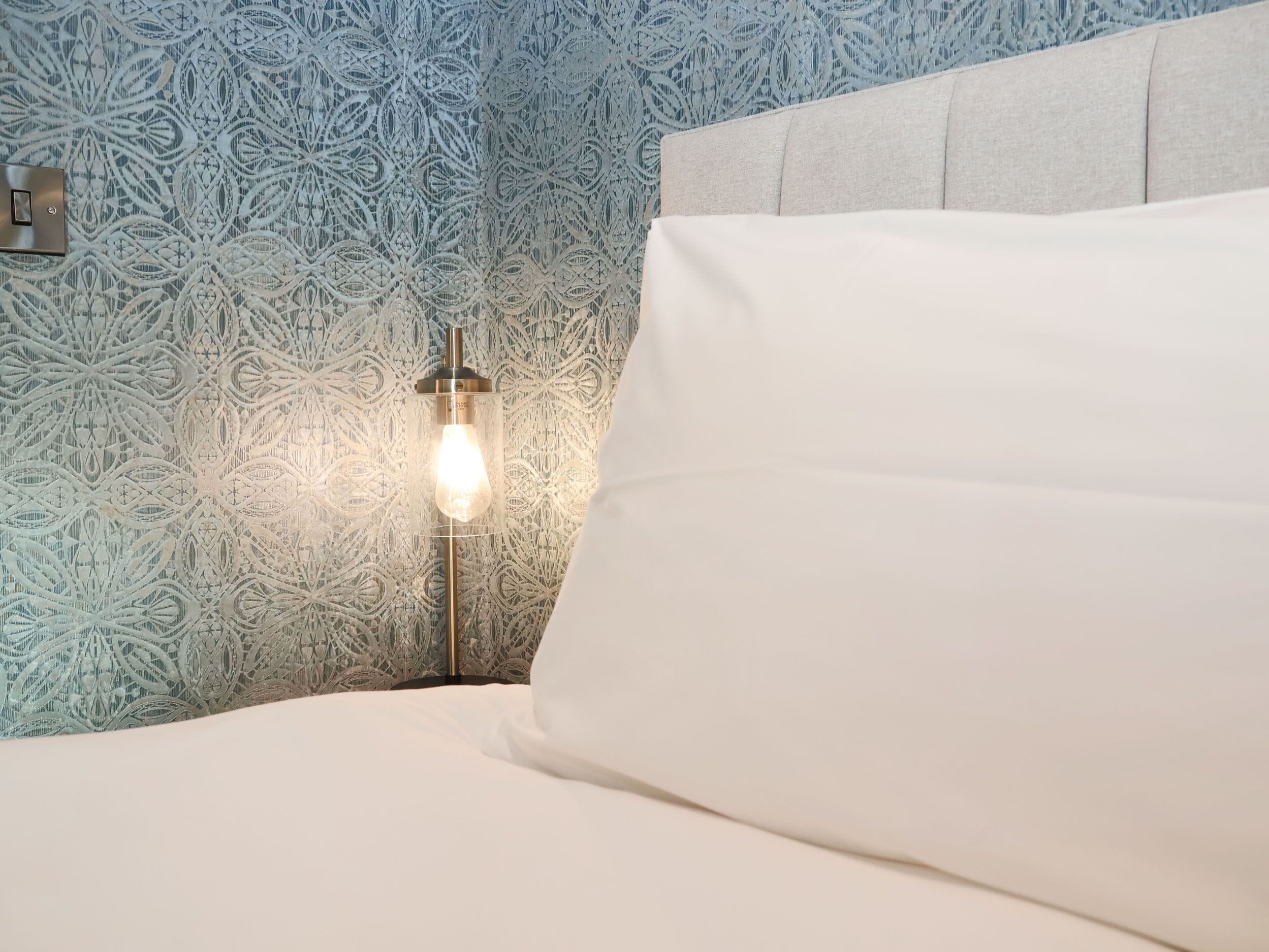 A bed with white bedding, a nightstand with a lamp, and a patterned wallpaper.