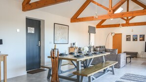 A spacious living area with a high ceiling and exposed wooden beams, a dining table set for six, a sofa, a bench, and a large window providing natural light. - The Stables (Lyme Regis)