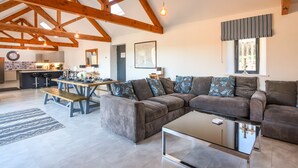 A spacious living area with a large sectional sofa, a glass coffee table, and a dining table set for four. The room features exposed wooden beams, a large window with a view, and a kitchen in the background. - The Stables (Lyme Regis)