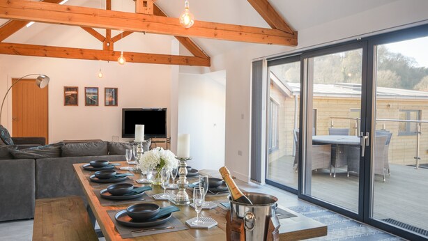 A dining area with a long wooden table set for a meal, a television in the background, and a view of the outside through large glass doors. - The Stables (Lyme Regis)