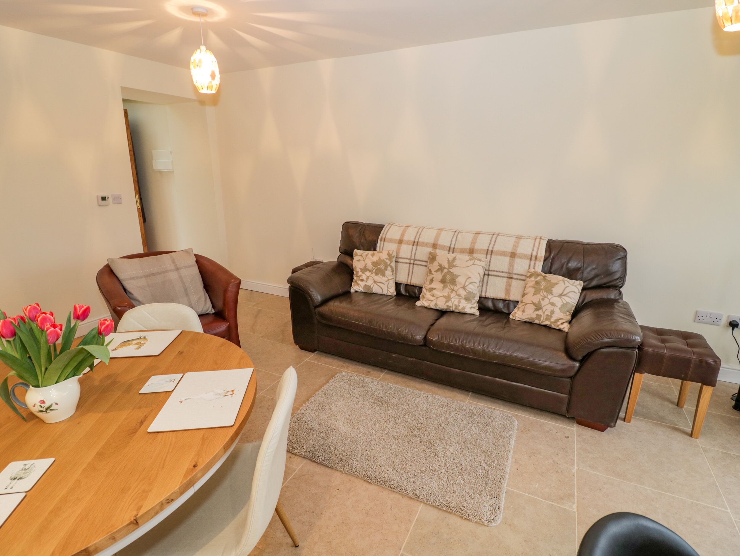 A living room with a brown leather sofa, a wooden dining table with tulips, and a white chair.