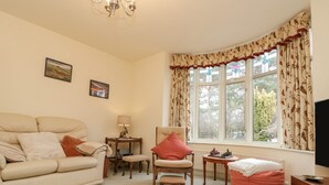 A living room with a sofa, armchair, ottoman, and a small table with a lamp. There is a window with curtains and a view of trees outside. - Clover Leaf (Guisborough)