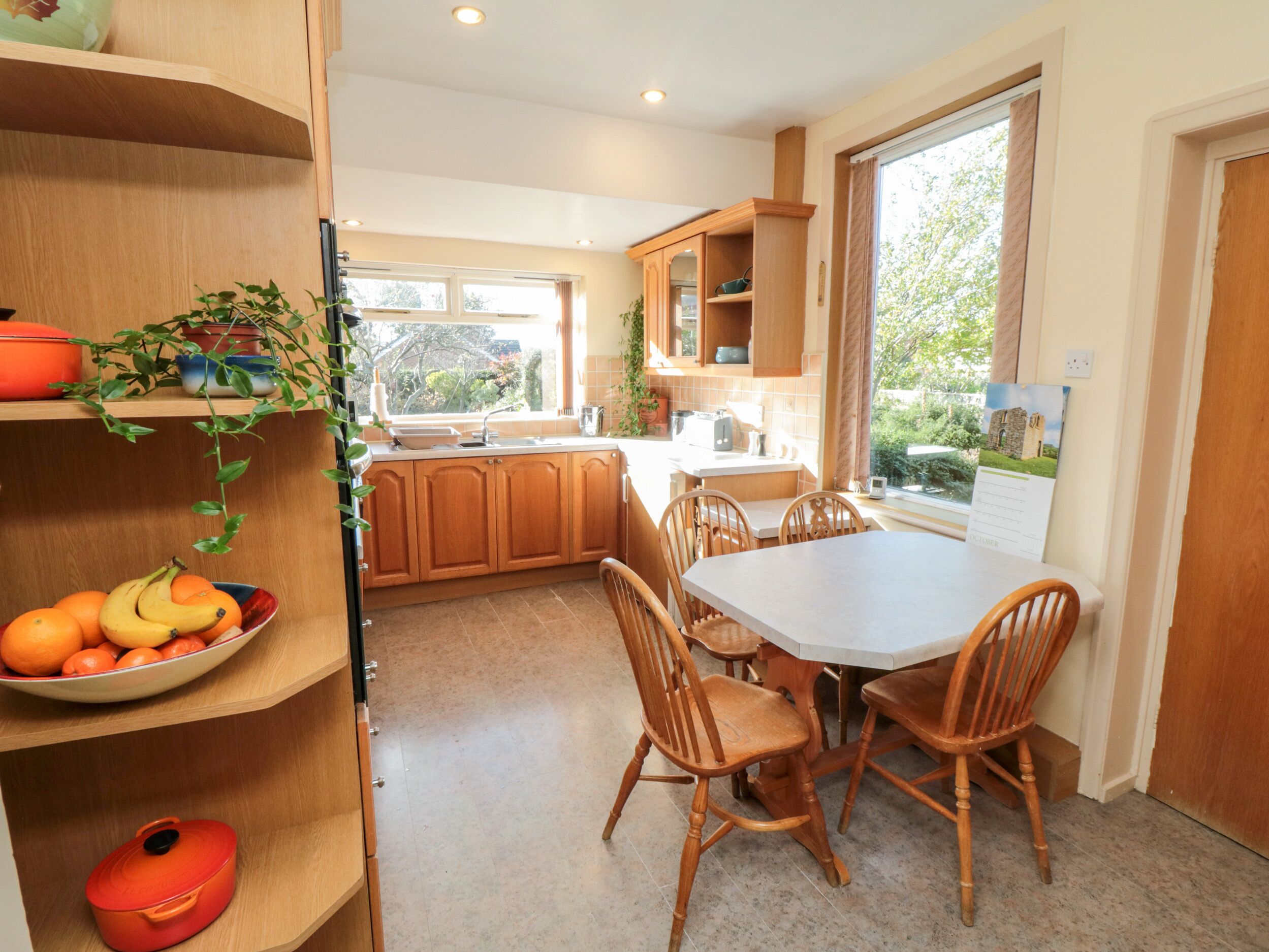 A kitchen with wooden cabinets, a dining table with chairs, a fruit bowl, and a window with a view of greenery.