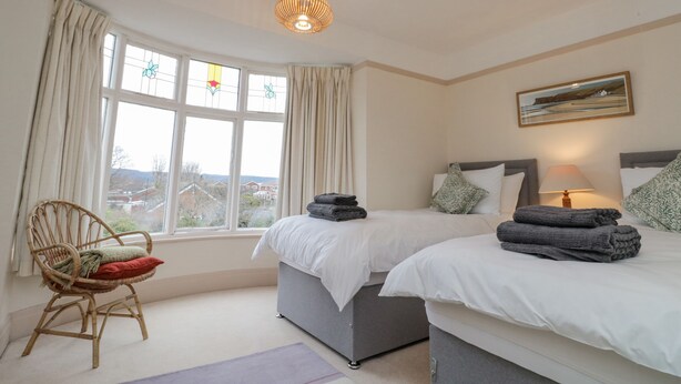 A bedroom with two beds, a wicker chair, a window with curtains, and a painting on the wall. - Clover Leaf (Guisborough)