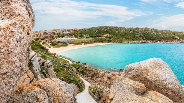 Vista costera con un camino pavimentado, terreno rocoso y edificios al fondo.