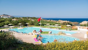 Una piscina con sillas de descanso y sombrillas, rodeada de vegetación y vista al mar.