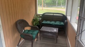 En lille veranda med fletmøbler, en potteplante og udsigt til en græsplæne og træer.