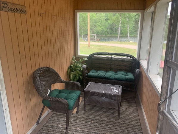 A small porch with wicker furniture, a potted plant, and a view of a grassy area and trees.