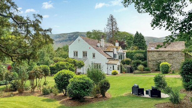 A white house with a tiled roof, surrounded by a well-maintained lawn and various shrubs.