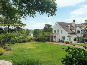 Une maison blanche avec un toit en pente, entourée d’une pelouse bien entretenue et de divers arbustes.