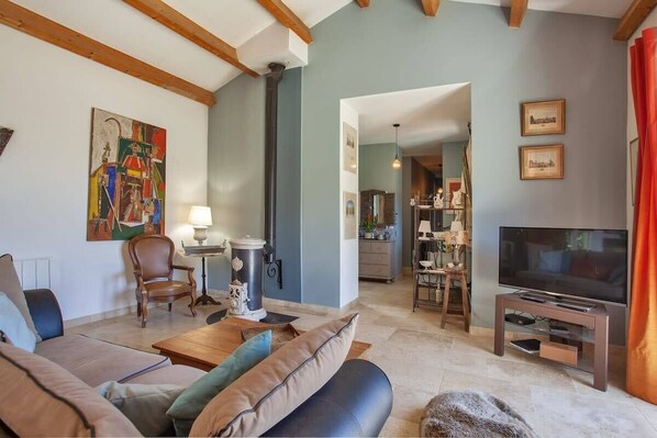 A living room with a sofa, armchair, and a television on a stand. There is a fireplace and a kitchen visible in the background. - Villa Merusellu (Sisco)