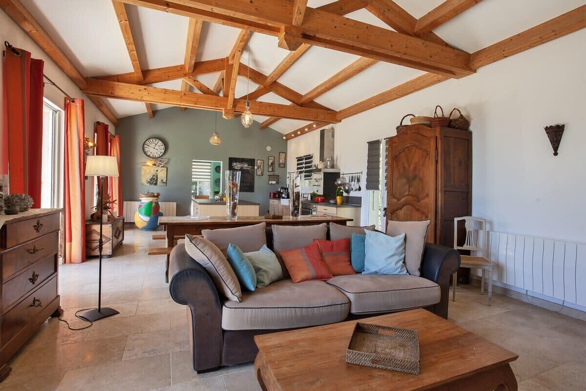 Un salon chaleureux avec un plafond en bois, un canapé agrémenté de coussins, une table basse et, au fond, une cuisine.