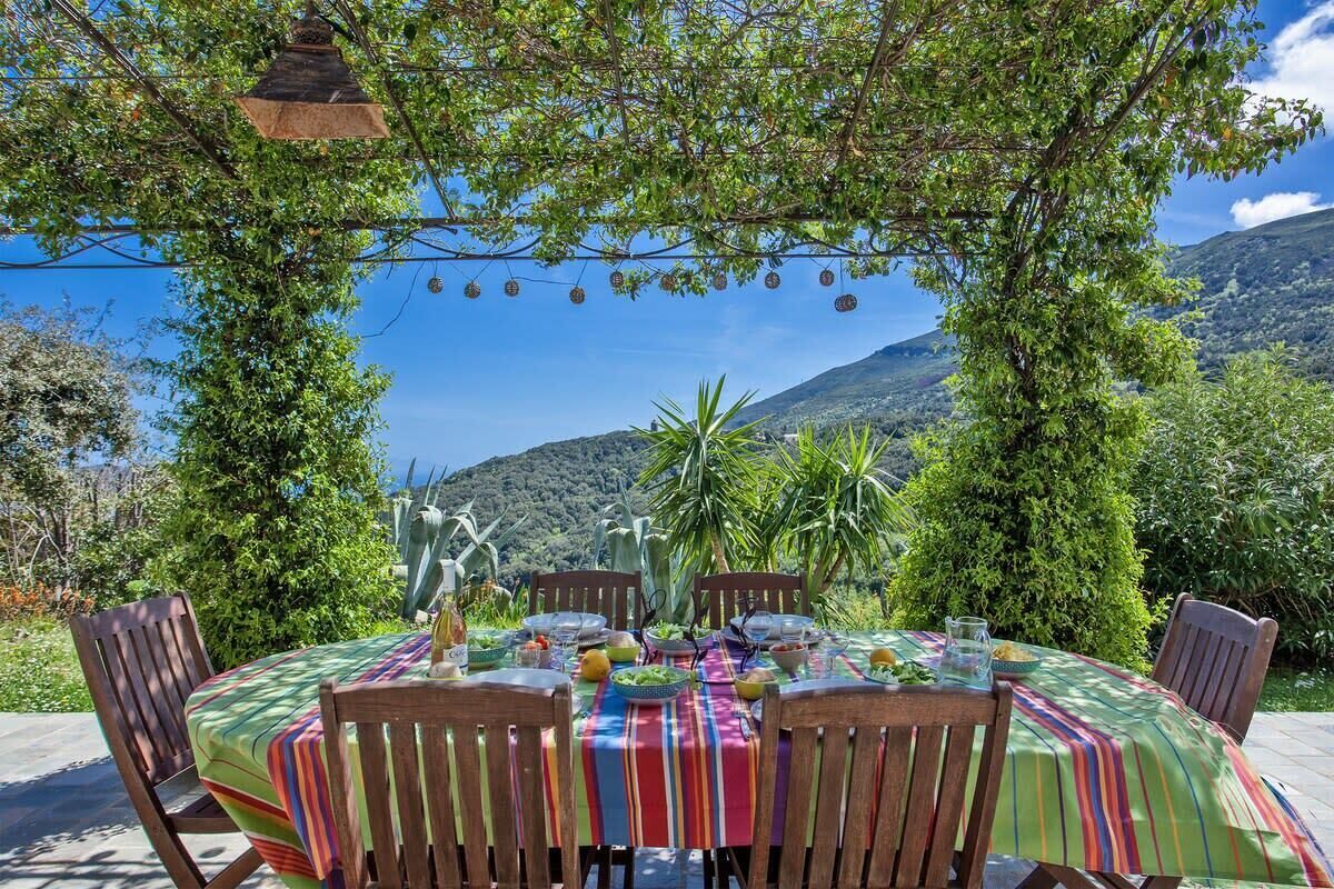 Un espace de restauration en plein air avec une table dressée pour quatre personnes, entourée de chaises en bois, sous une pergola agrémentée de décorations suspendues.